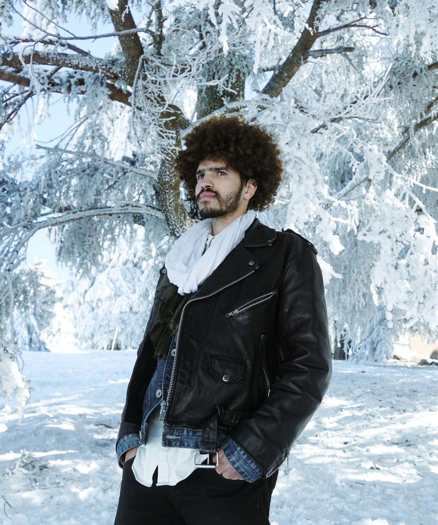 Young man with afro hair wearing black leather jacket standing in snowy winter landscape with frost-covered trees.