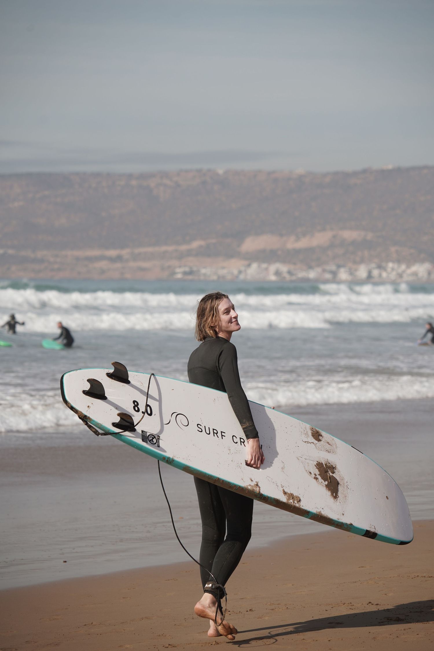 A surfer in a wetsuit carries a white surfboard along a sandy beach with ocean waves in the background.