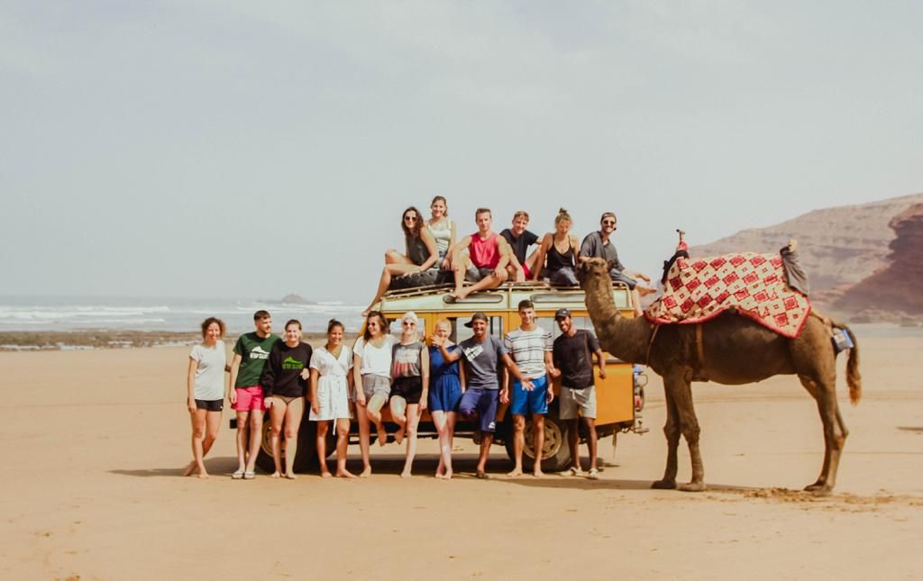 Group of tourists posing on beach with camel and jeep in Morocco desert landscape on sunny day.