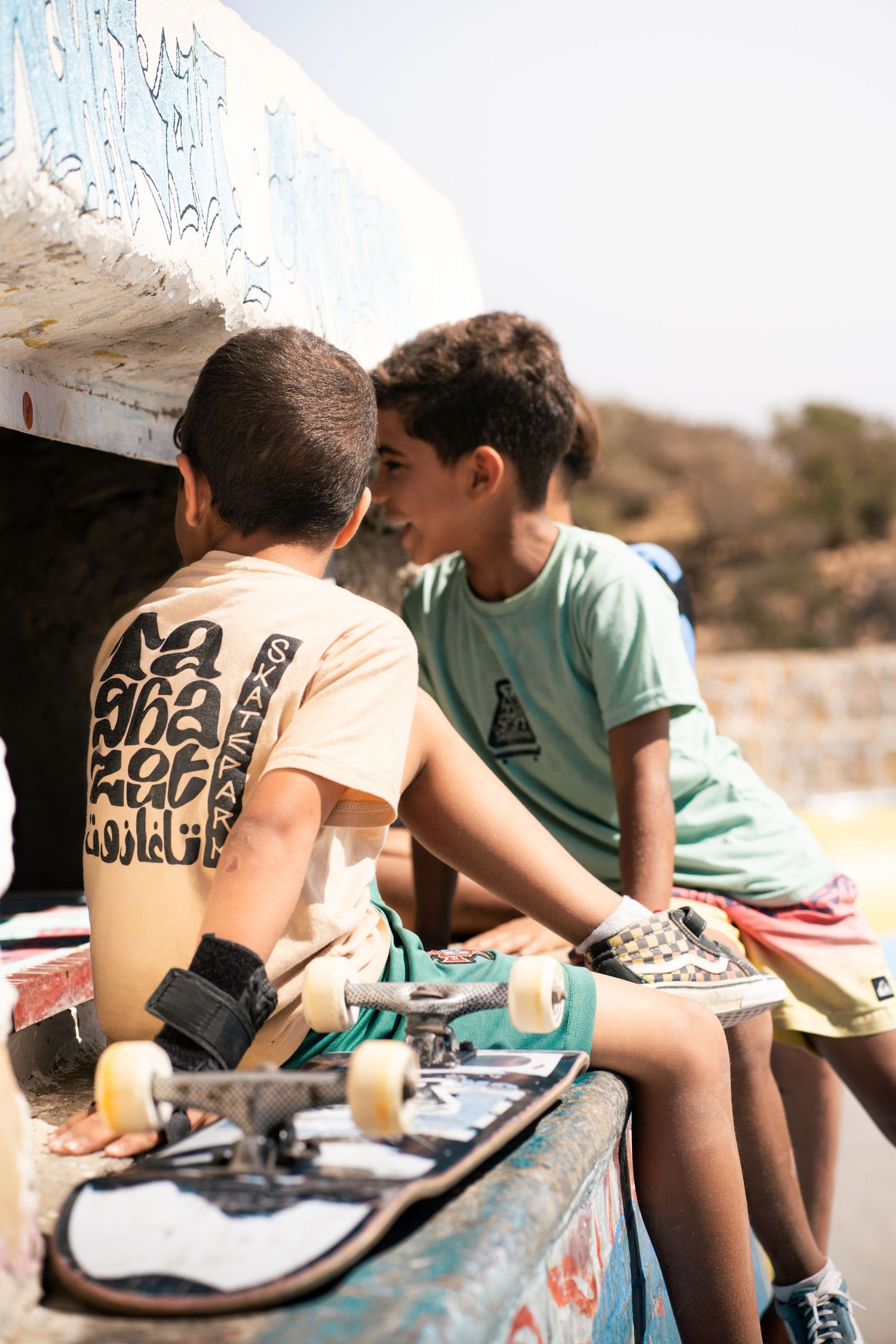 Two young boys with skateboards sitting together at an outdoor skate park on a sunny day.