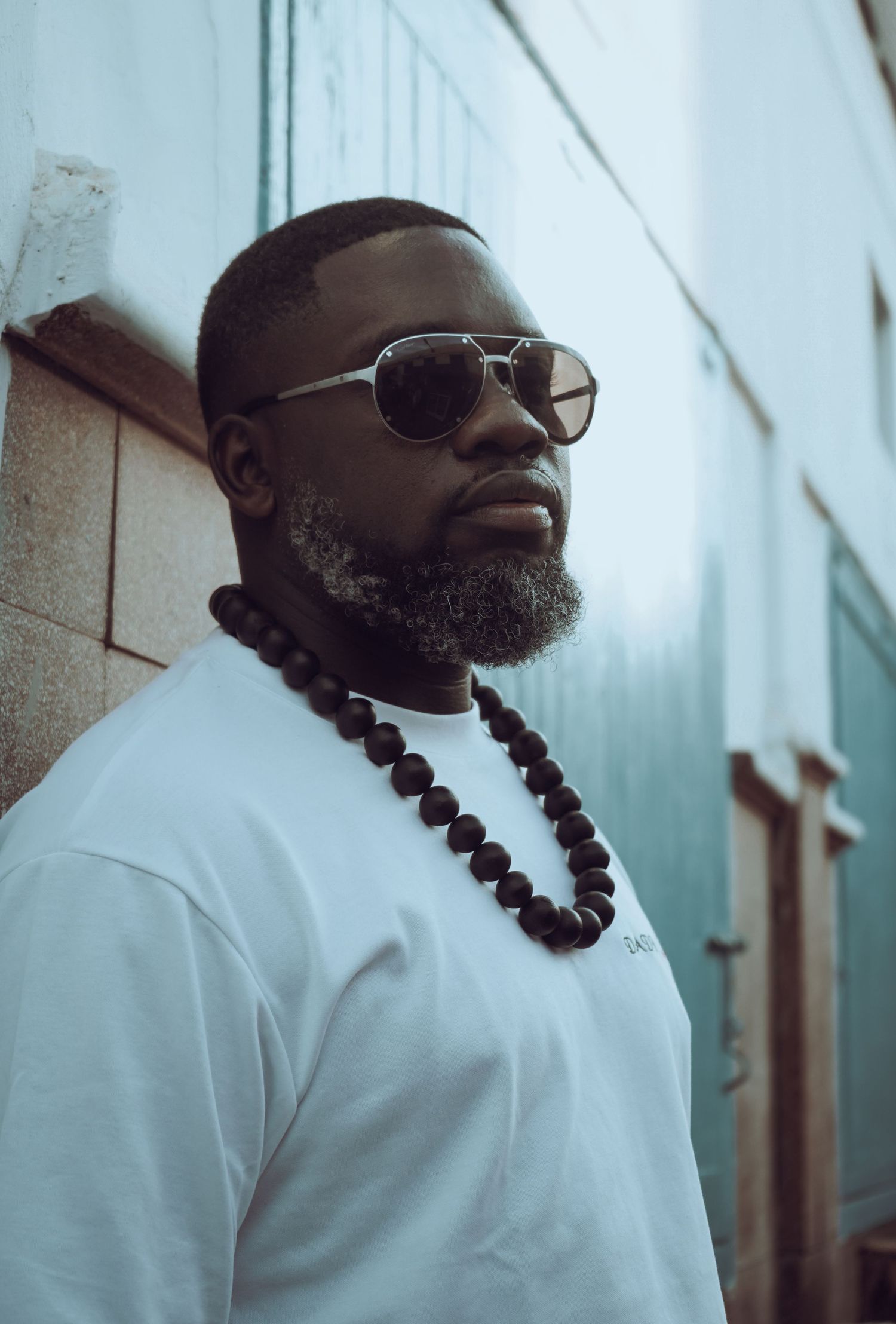 Stylish Black man with beard wearing sunglasses, white tee, and large black beaded necklace outdoors.
