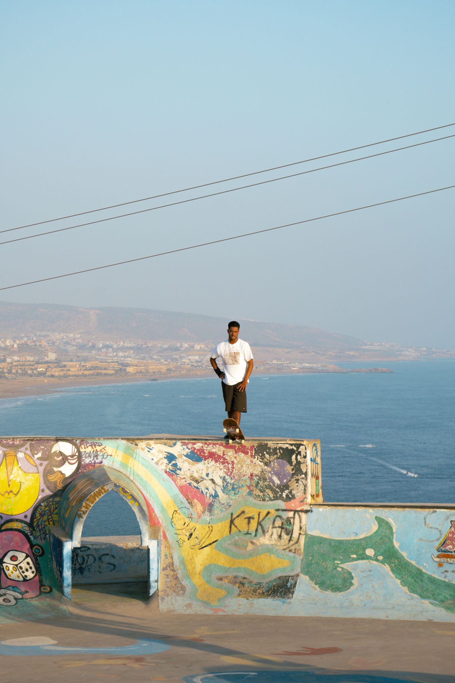 Skateboarder standing on a graffiti-covered ramp at a skate park overlooking the ocean coastline.