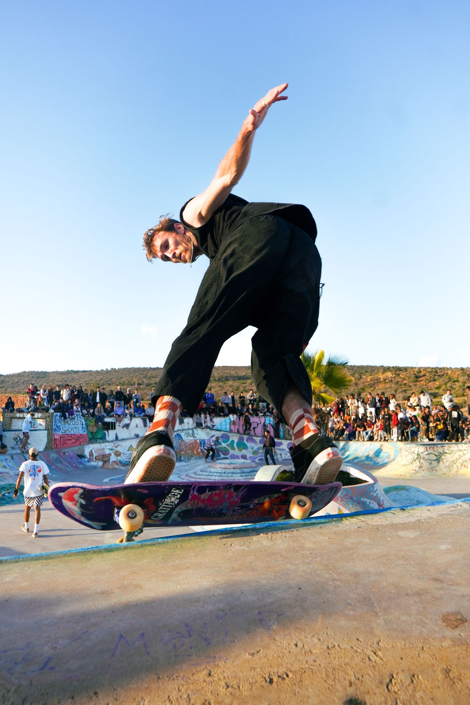 Skateboarder executing nose grind trick on concrete ledge with large crowd spectating at outdoor skatepark.