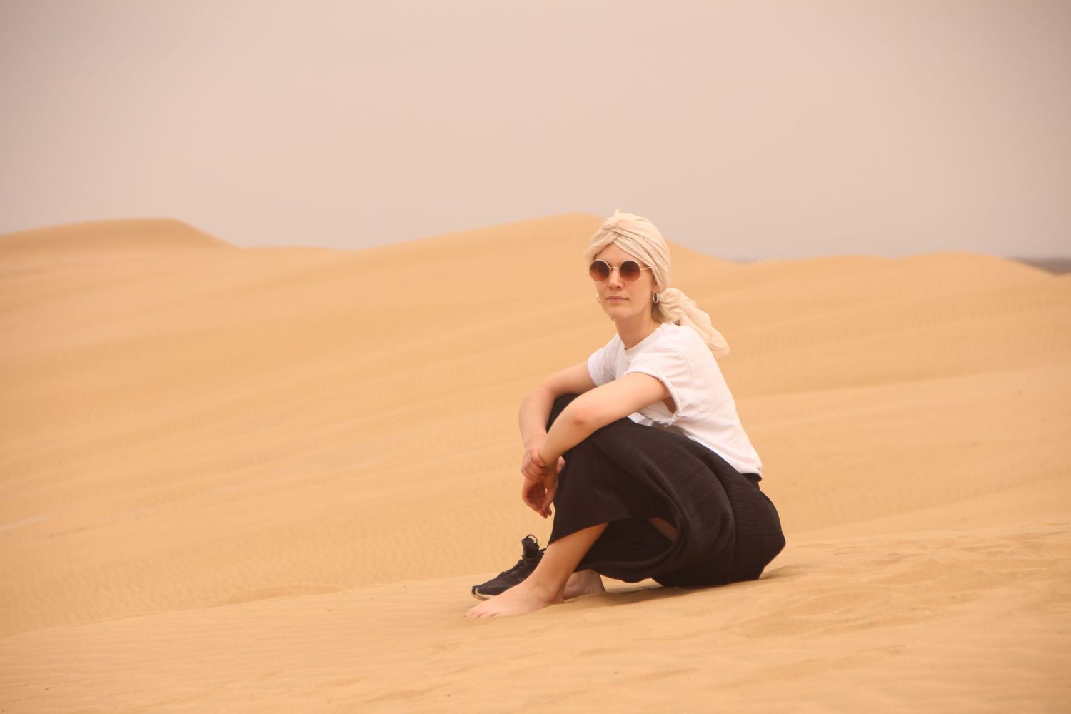 Blonde woman in sunglasses sitting on golden sand dunes in the desert, wearing white top and black skirt.