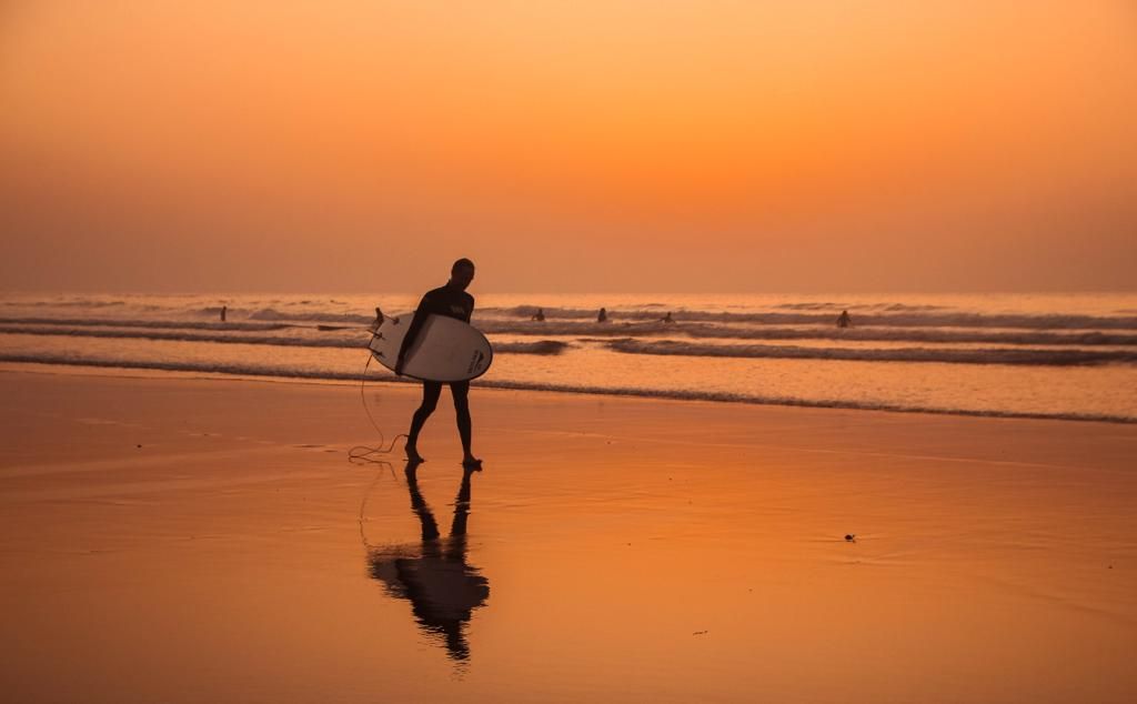Silhouette of a surfer carrying a white surfboard walking on a reflective beach during a stunning golden sunset.
