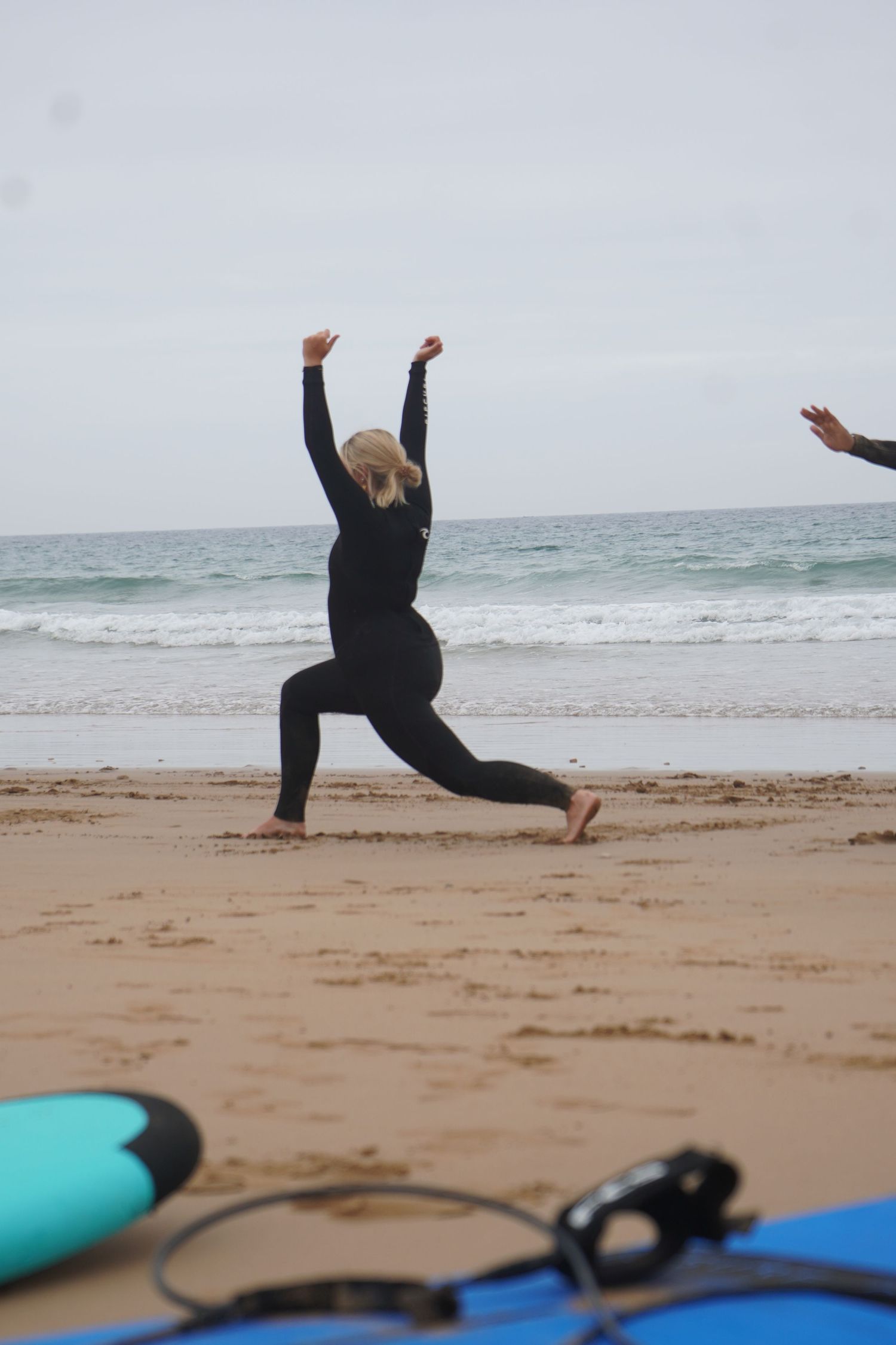 Woman in black wetsuit doing a yoga lunge pose on sandy beach with surfboards and ocean waves in background.