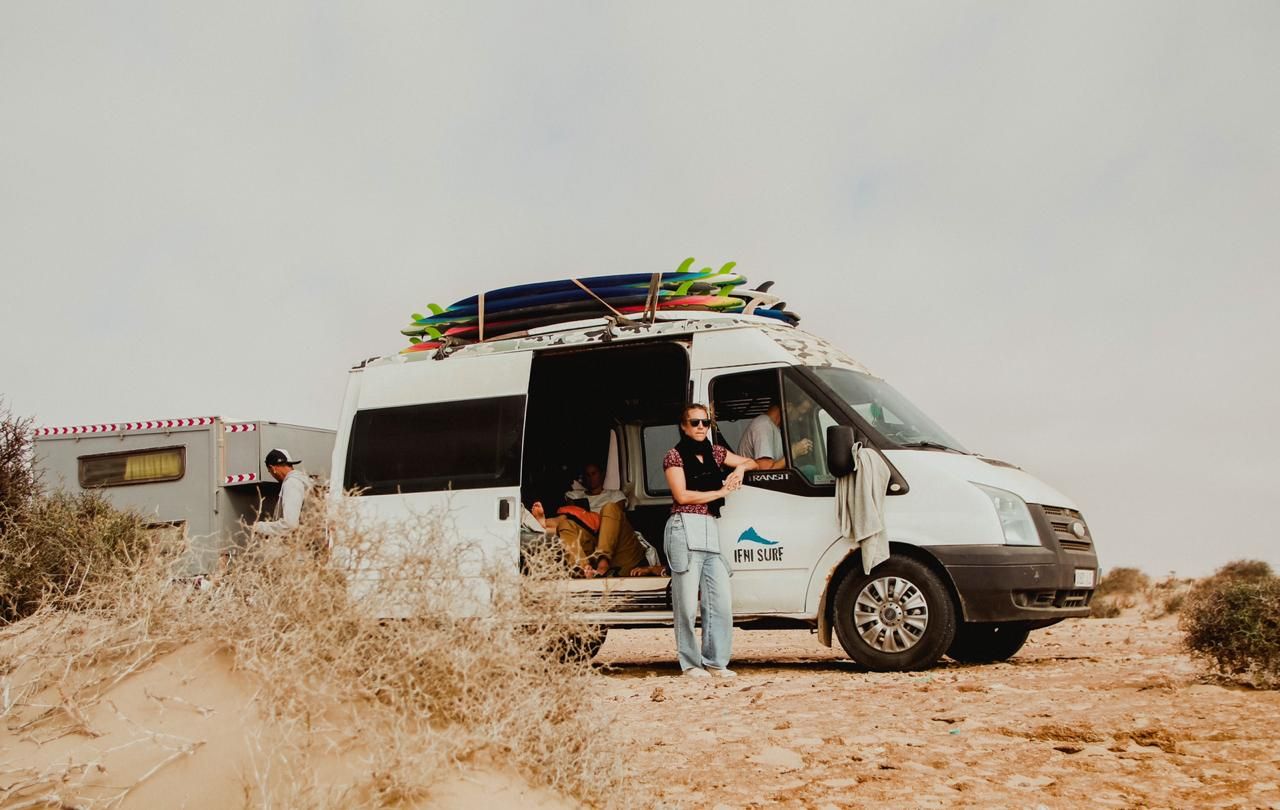Man standing beside a Ford Transit camper van with surfboards on roof rack in a sandy desert landscape.