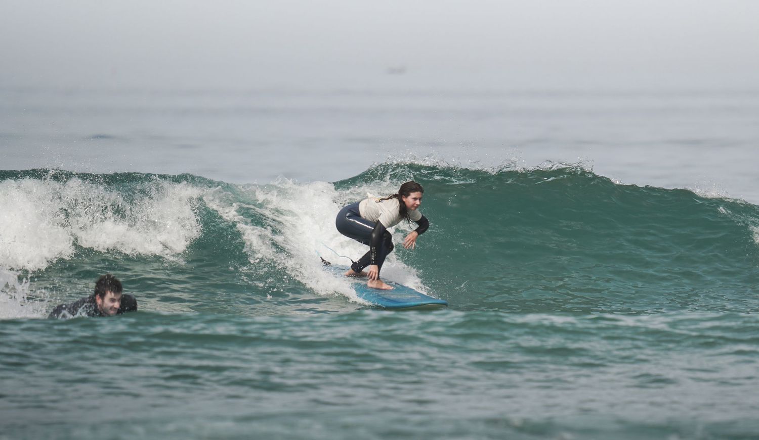 A surfer rides a wave on a blue surfboard while another person swims nearby in the ocean.