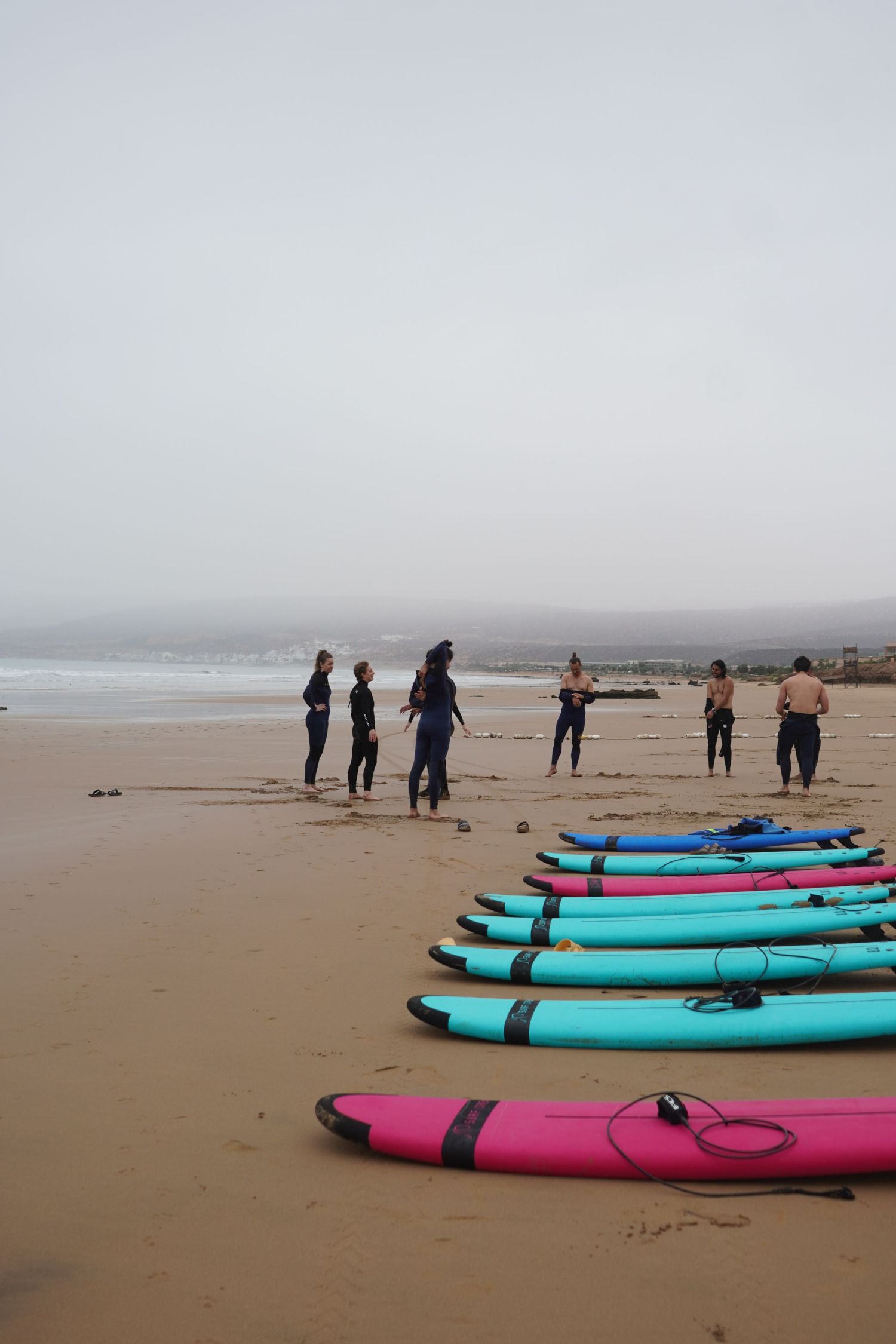 Colorful pink and blue surfboards lined up on a sandy beach with a group of surfers receiving lessons on a foggy day.