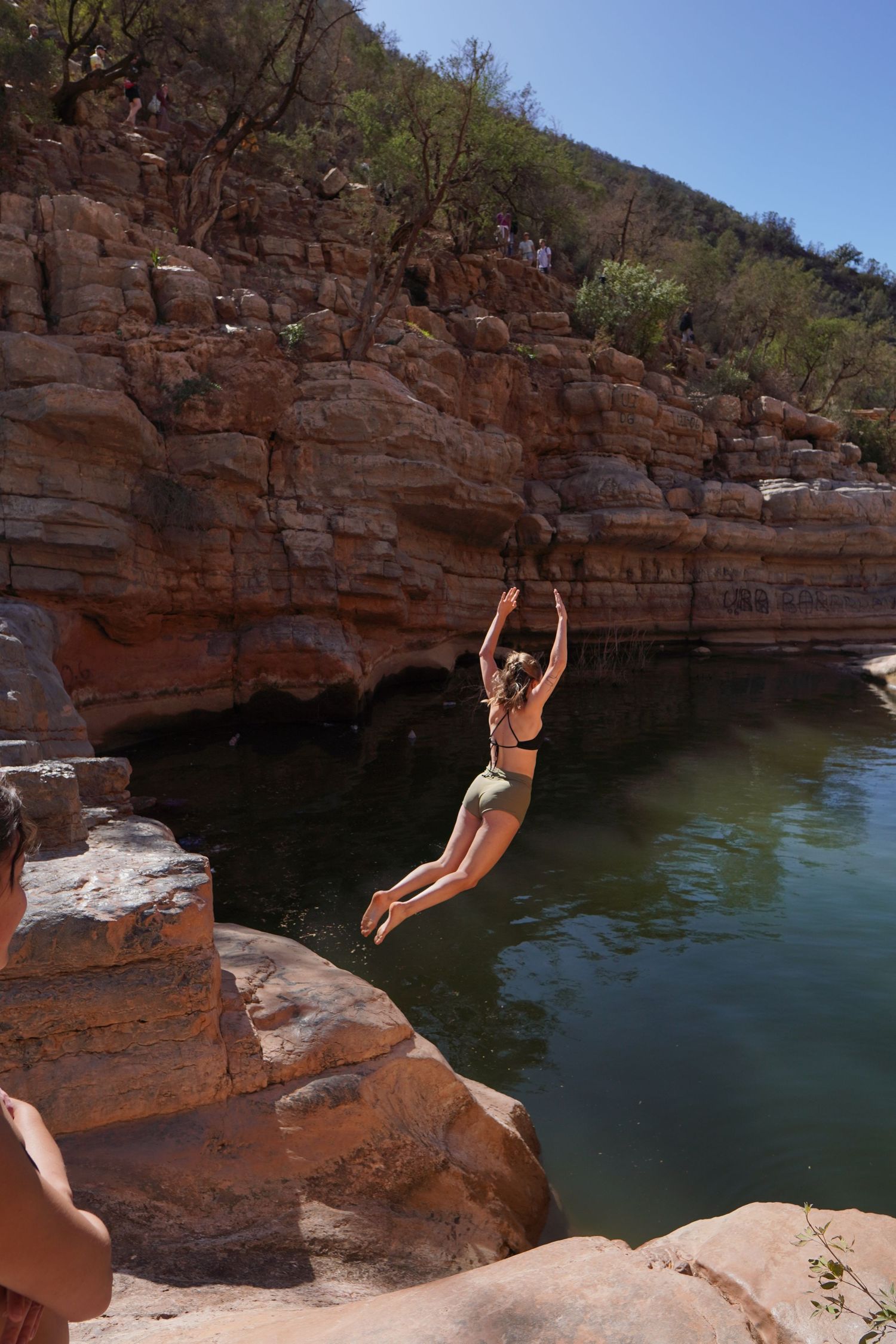Woman in bikini cliff jumping into a natural rock pool surrounded by red sandstone cliffs on a sunny day.