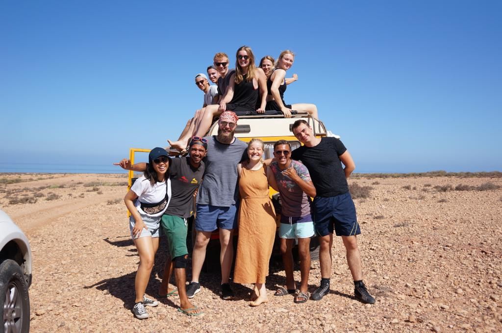 Group of friends posing together on a desert road trip, gathered around a yellow vehicle under clear blue skies.