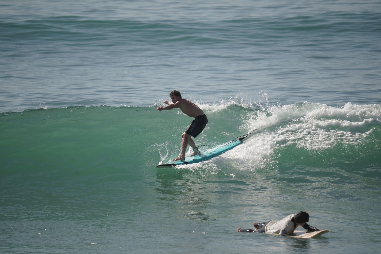 A surfer rides a wave on a blue surfboard in the ocean, with another surfer lying on a board nearby.
