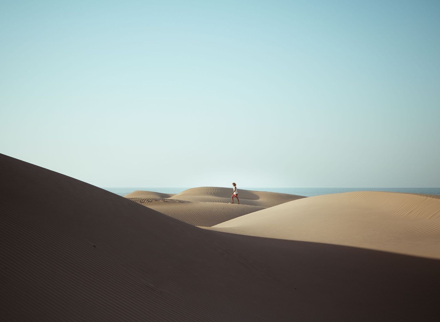A lone figure stands atop a vast golden sand dune under a clear blue sky in a serene desert landscape.