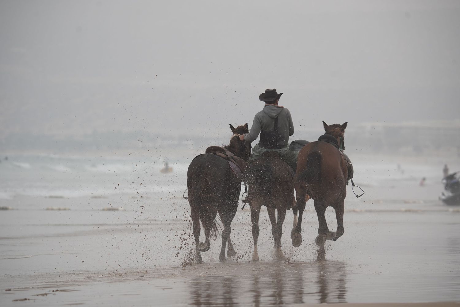 A cowboy rides horses along a misty beach shoreline with waves in the background.