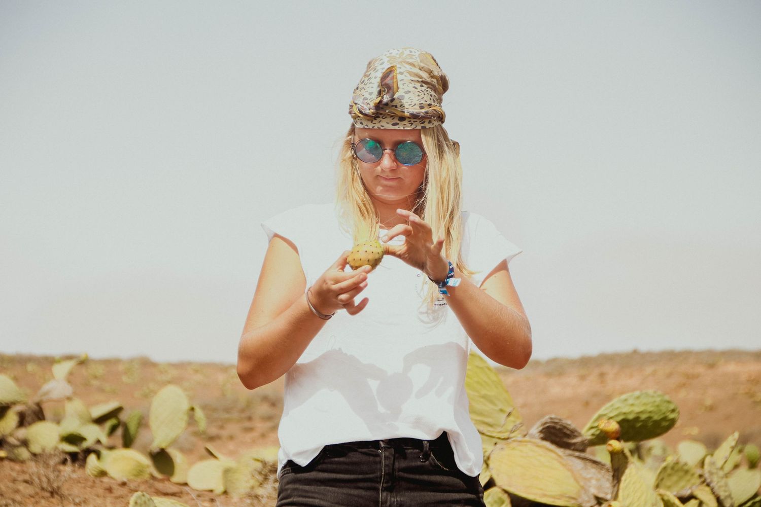 Blonde woman in white tee and head scarf eating cactus fruit in a desert landscape.