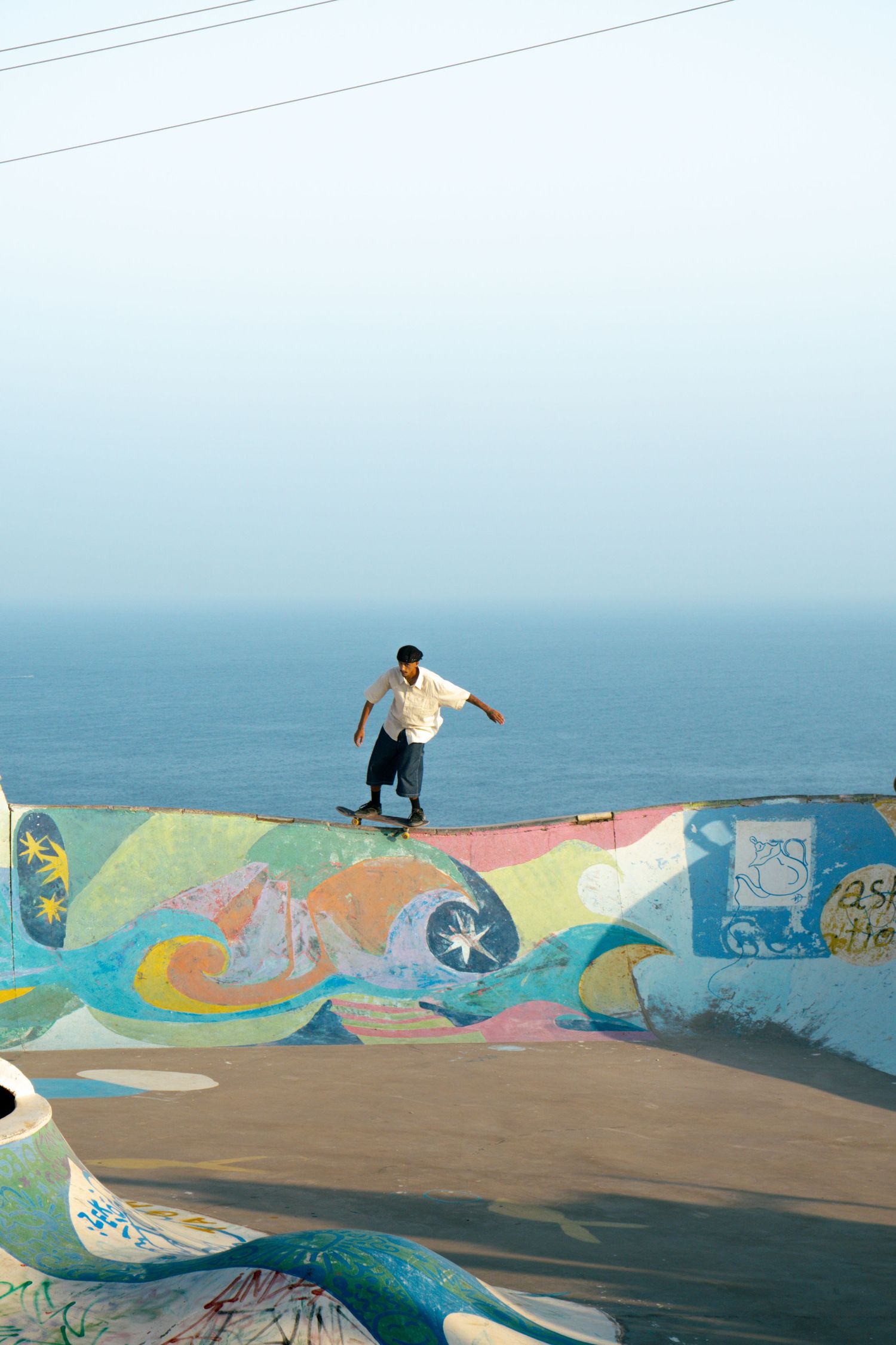 Skateboarder riding colorful mural-painted ramp wall with ocean horizon visible in background at coastal skatepark.
