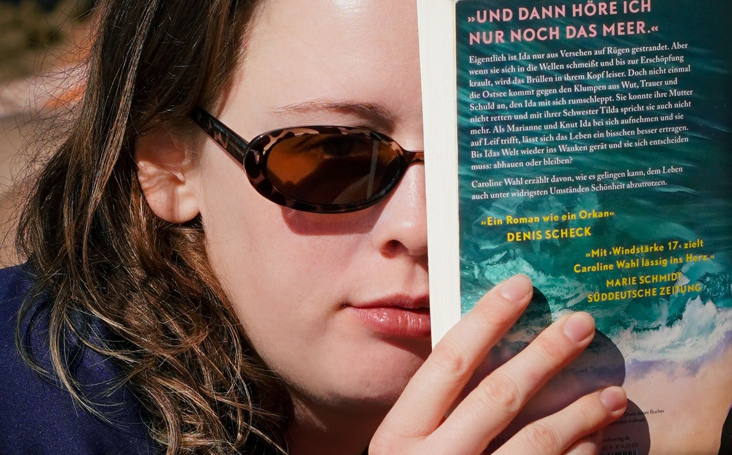 Woman in sunglasses reading a German book outdoors, holding it close to her face.