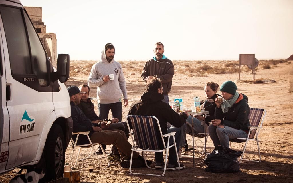 Group of people gathered around chairs in a desert landscape near a white van, enjoying an outdoor meeting.