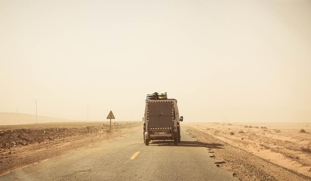 A van drives along a desert highway through a sandy haze, with rocky terrain and a warning sign visible nearby.