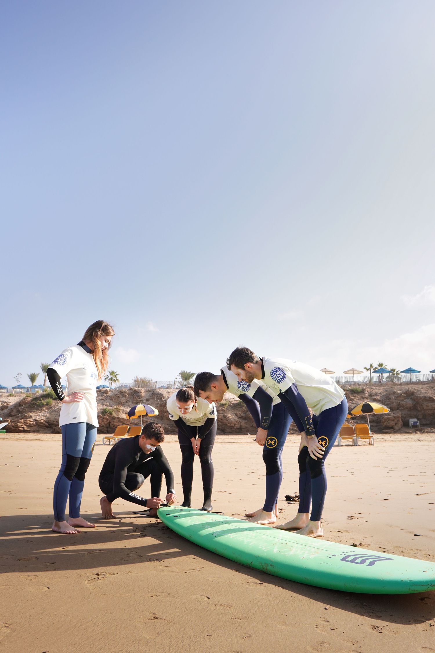 Group of surfers in wetsuits examining a green surfboard on a sandy beach during a surf lesson.