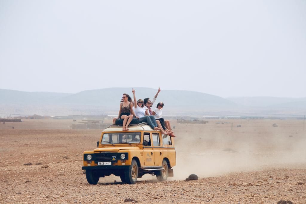 Group of friends riding on top of a yellow Land Rover through a vast desert landscape, raising arms in excitement.