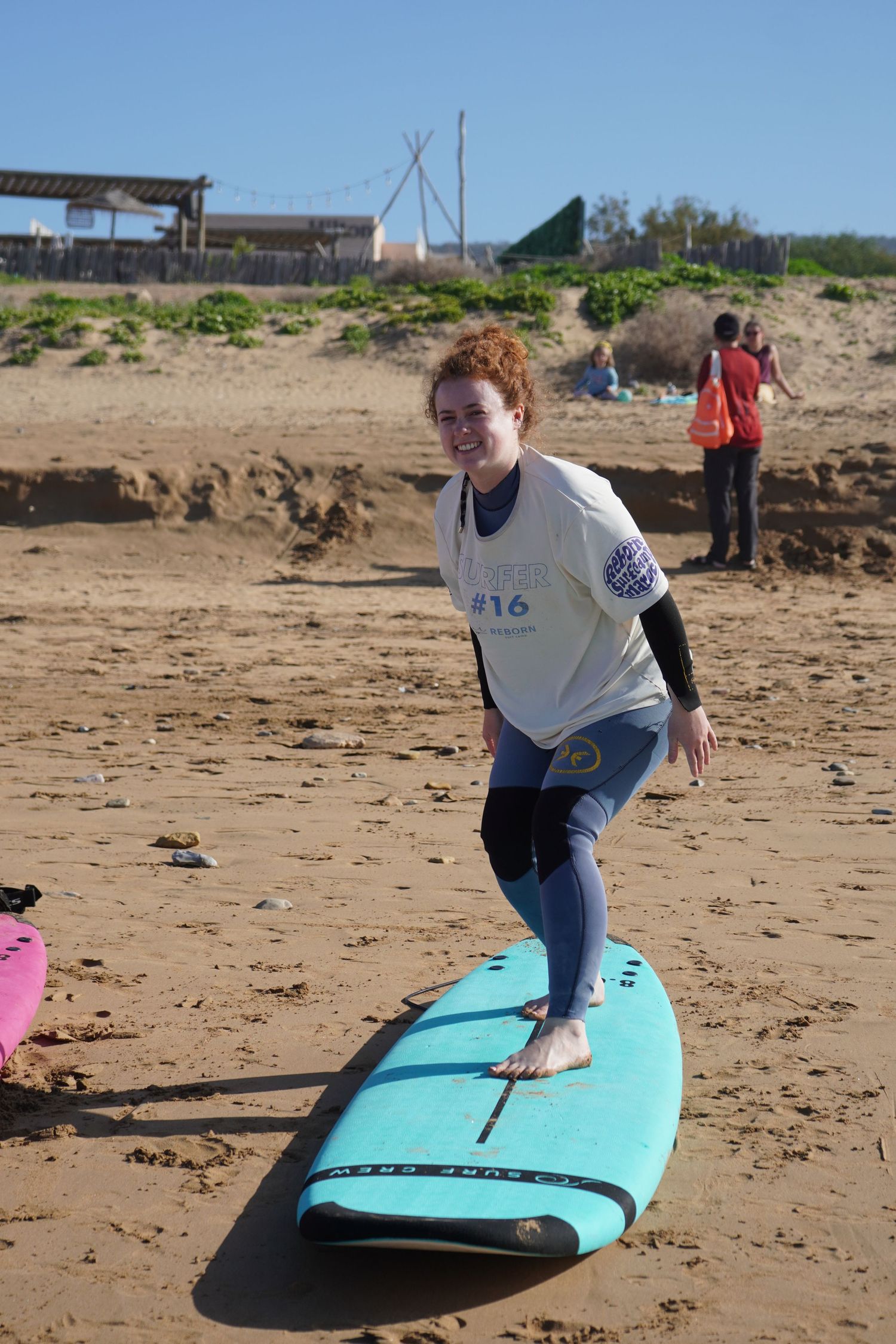 Young woman in wetsuit practicing surfing stance on blue surfboard on sandy beach during surf lesson.