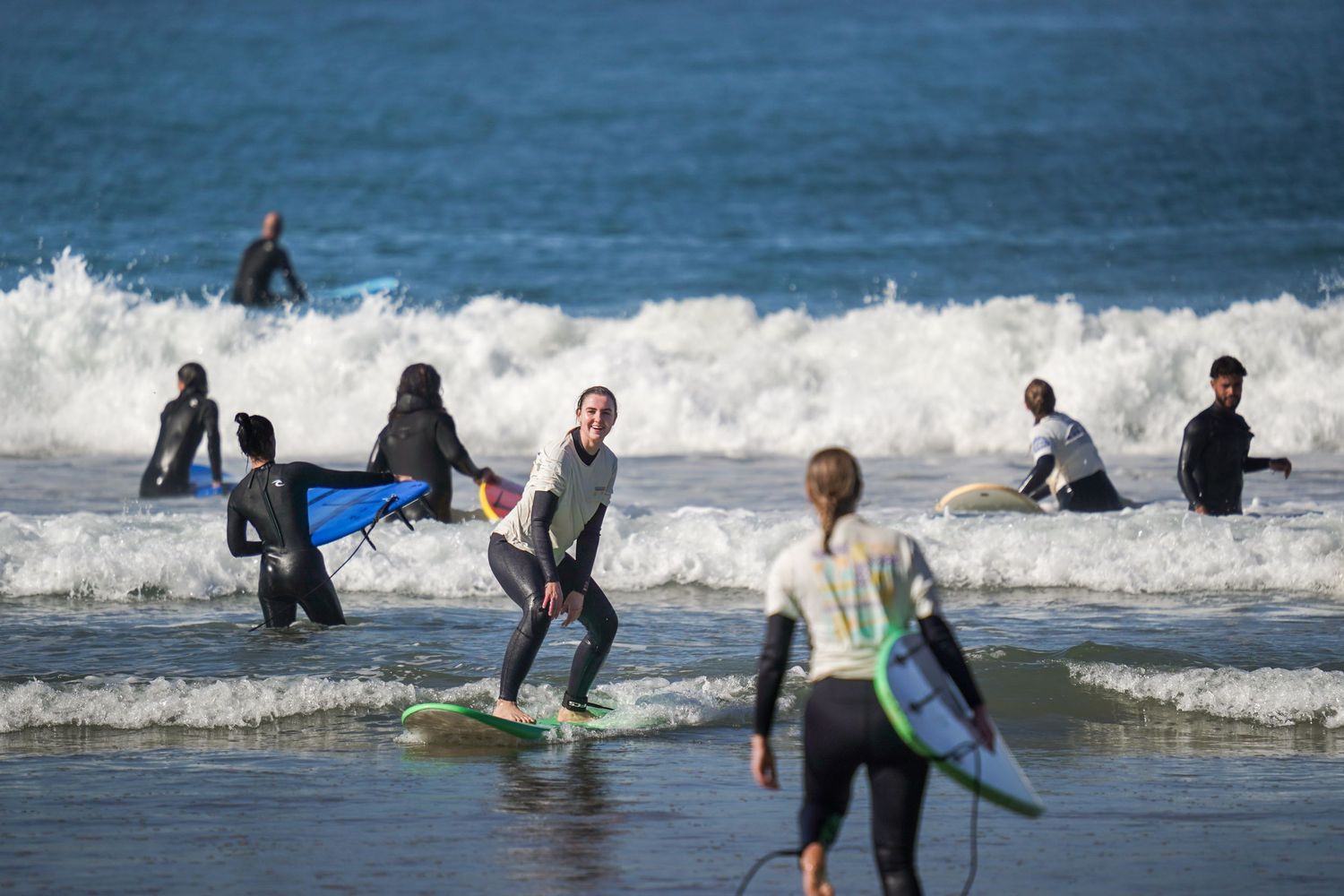 Group of surfers in wetsuits carrying boards and riding waves on a sunny beach day.