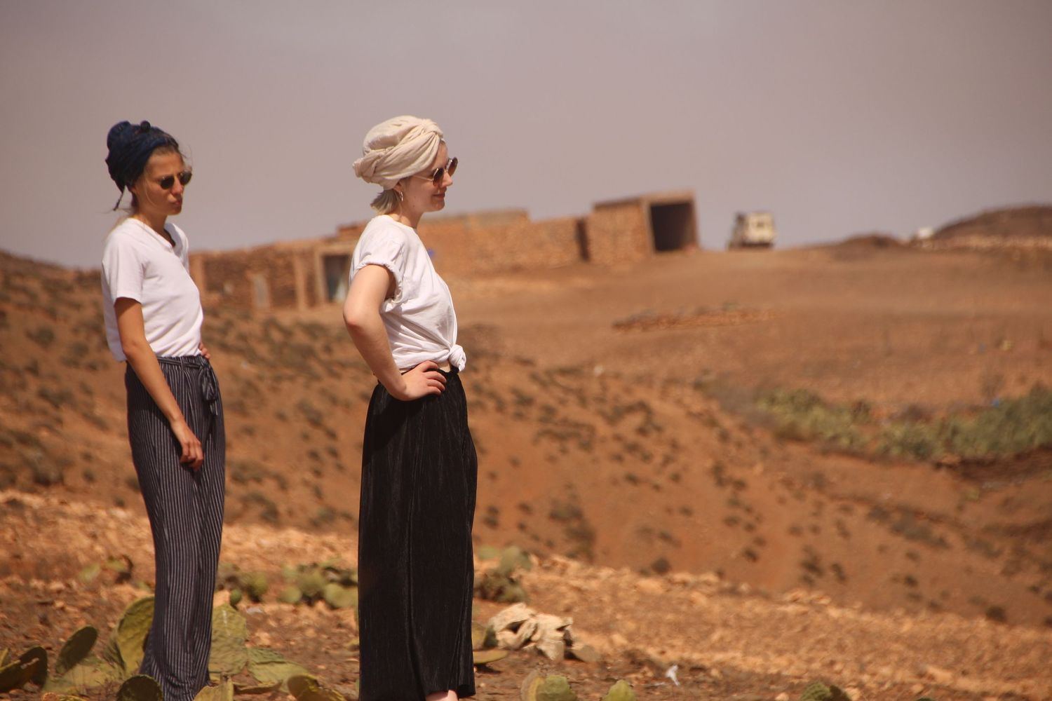 Two women in white tops and dark pants stand on a red dirt hillside with ancient ruins in an arid landscape.