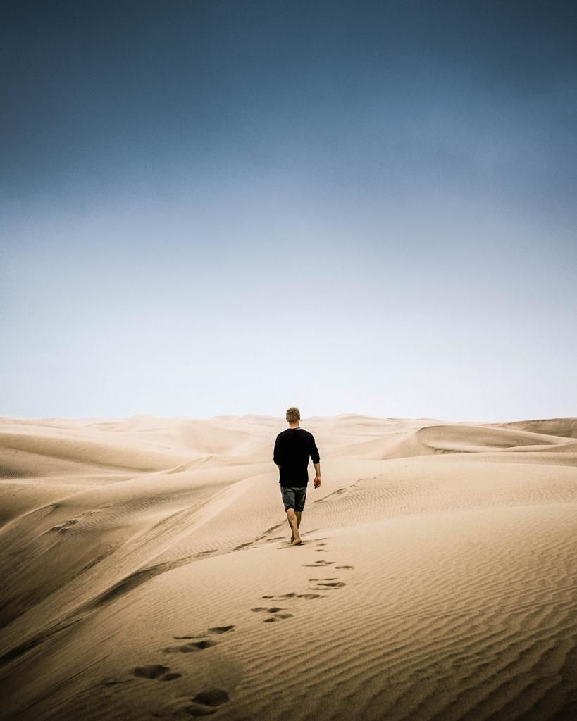 A lone person walks across vast golden sand dunes leaving footprints behind under a clear blue sky.