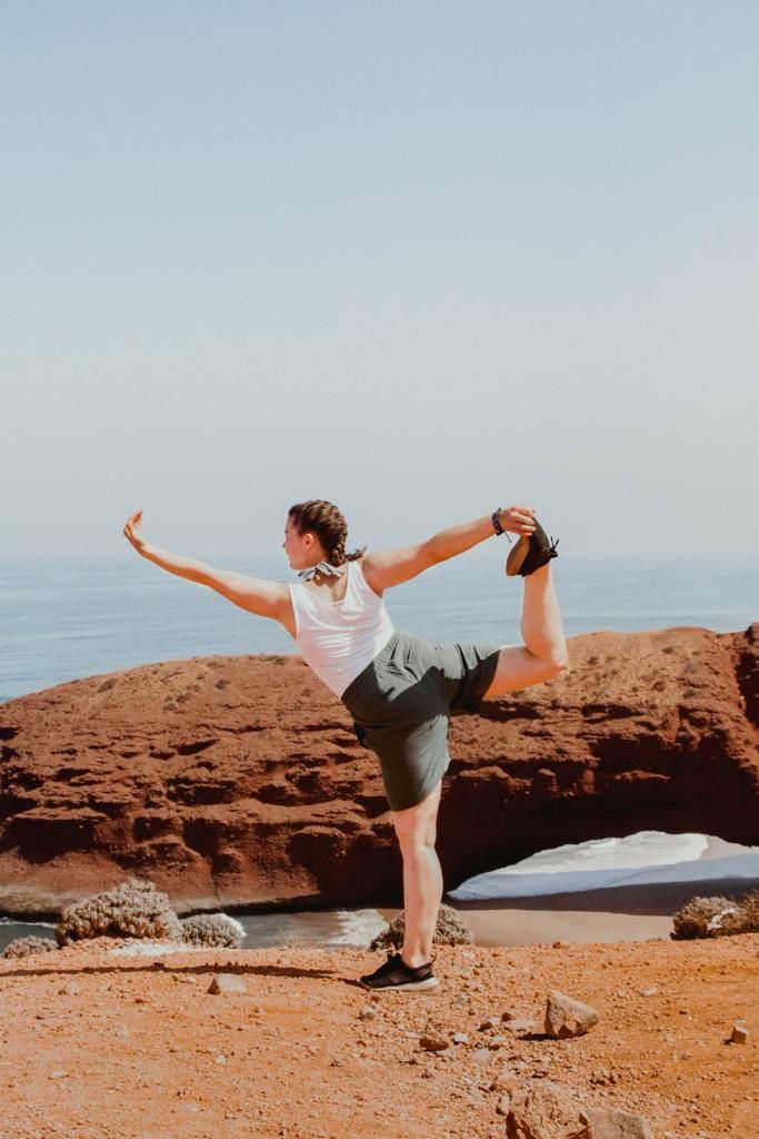 Woman practicing yoga dancer pose on red rock cliff overlooking the ocean on a sunny day.
