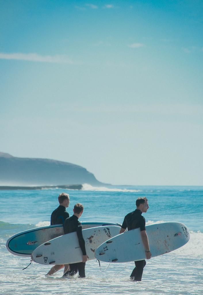 Two surfers in wetsuits carry surfboards through ocean waves on a sunny beach with a rocky headland.
