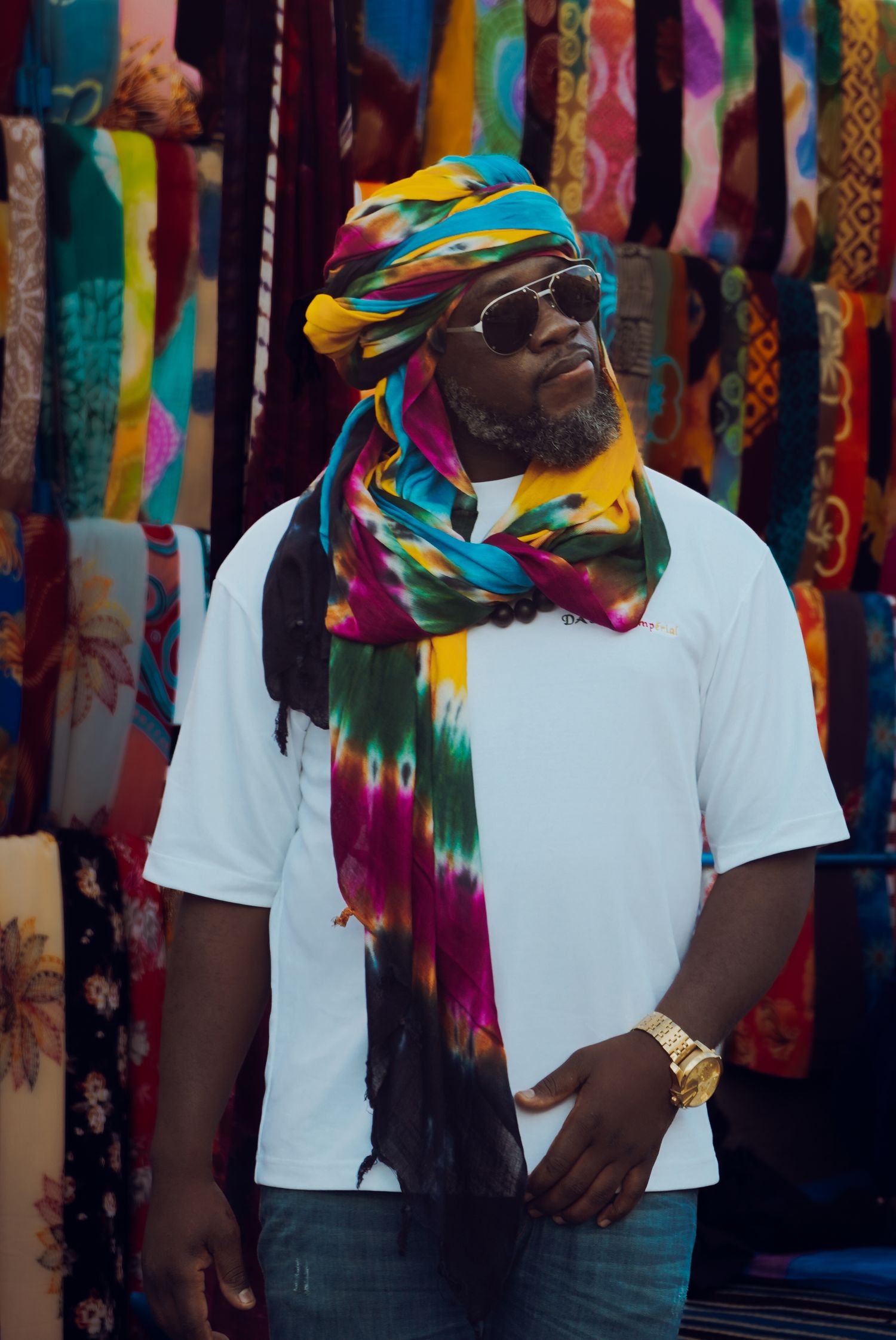 Man wearing colorful tie-dye turban and white t-shirt posing in front of vibrant textile market stall.