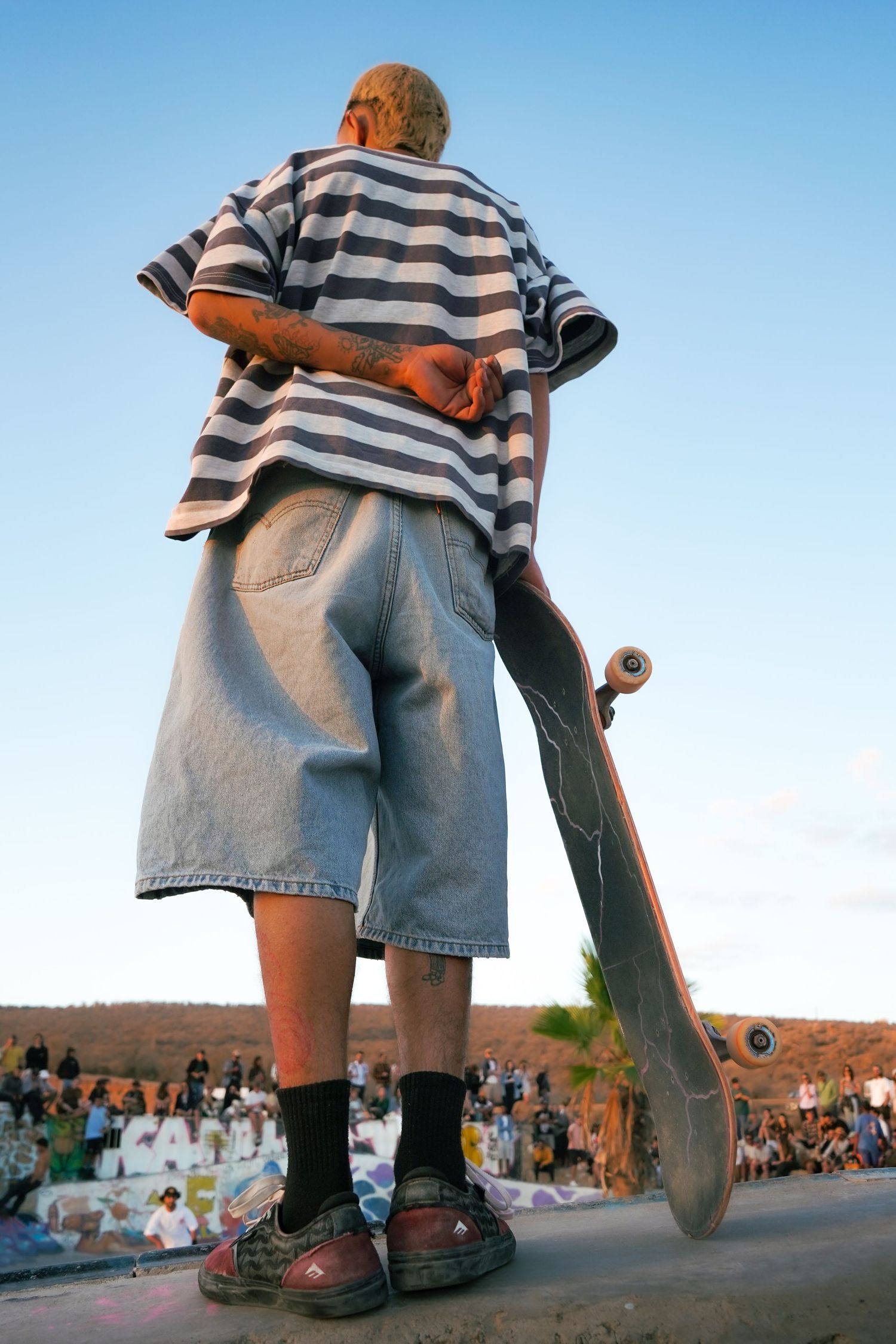 Rear view of tattooed skateboarder holding board upright at edge of ramp before a large crowd at sunset.