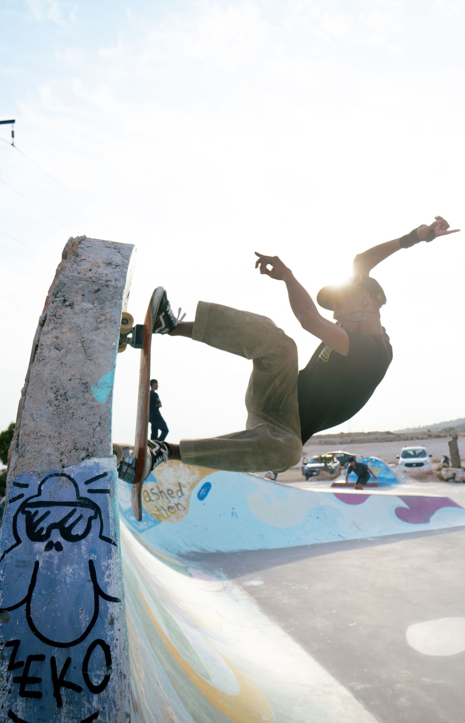 Skateboarder catching air on ramp at graffiti-decorated outdoor skate park in bright sunlight.