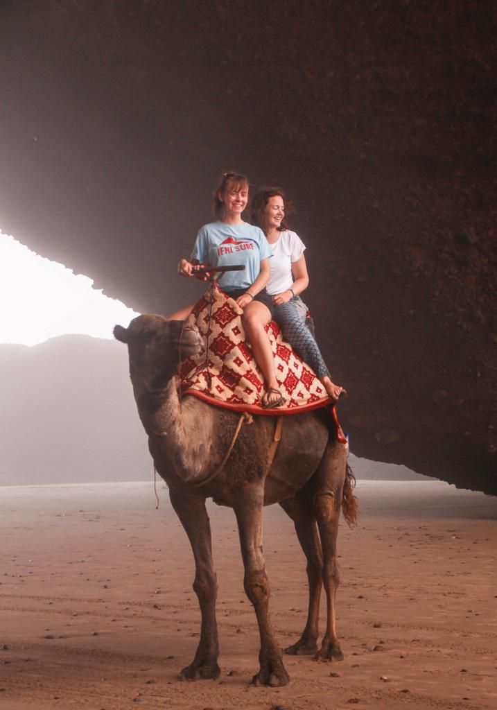 Two women riding a camel through a sandy desert landscape near a large rock formation.