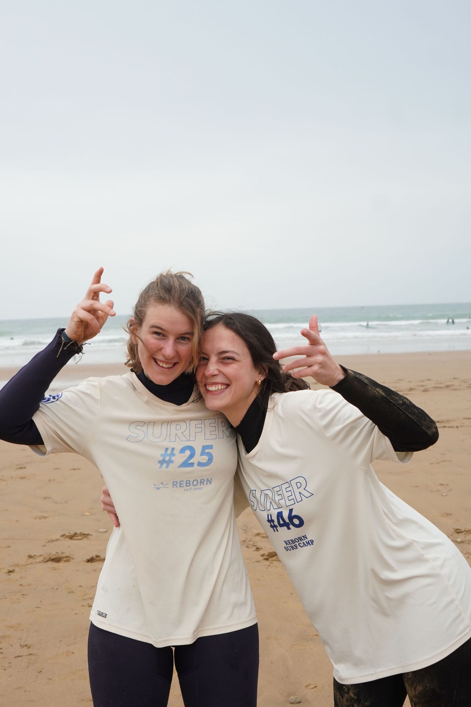 Two smiling surfers in numbered competition shirts pose together on a sandy beach with ocean waves in the background.