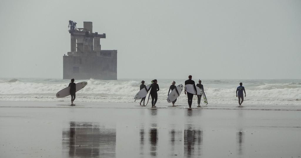 Group of surfers walking on a misty beach toward the ocean with a large industrial structure in the background.
