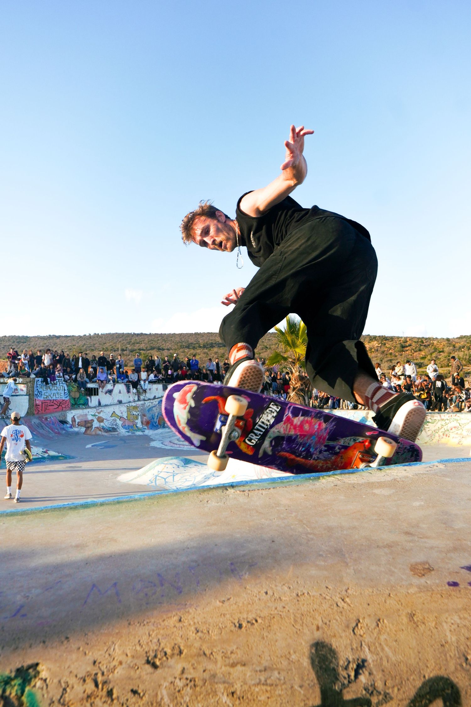 Skateboarder performing aerial trick at outdoor skatepark with crowd watching in background under blue sky.