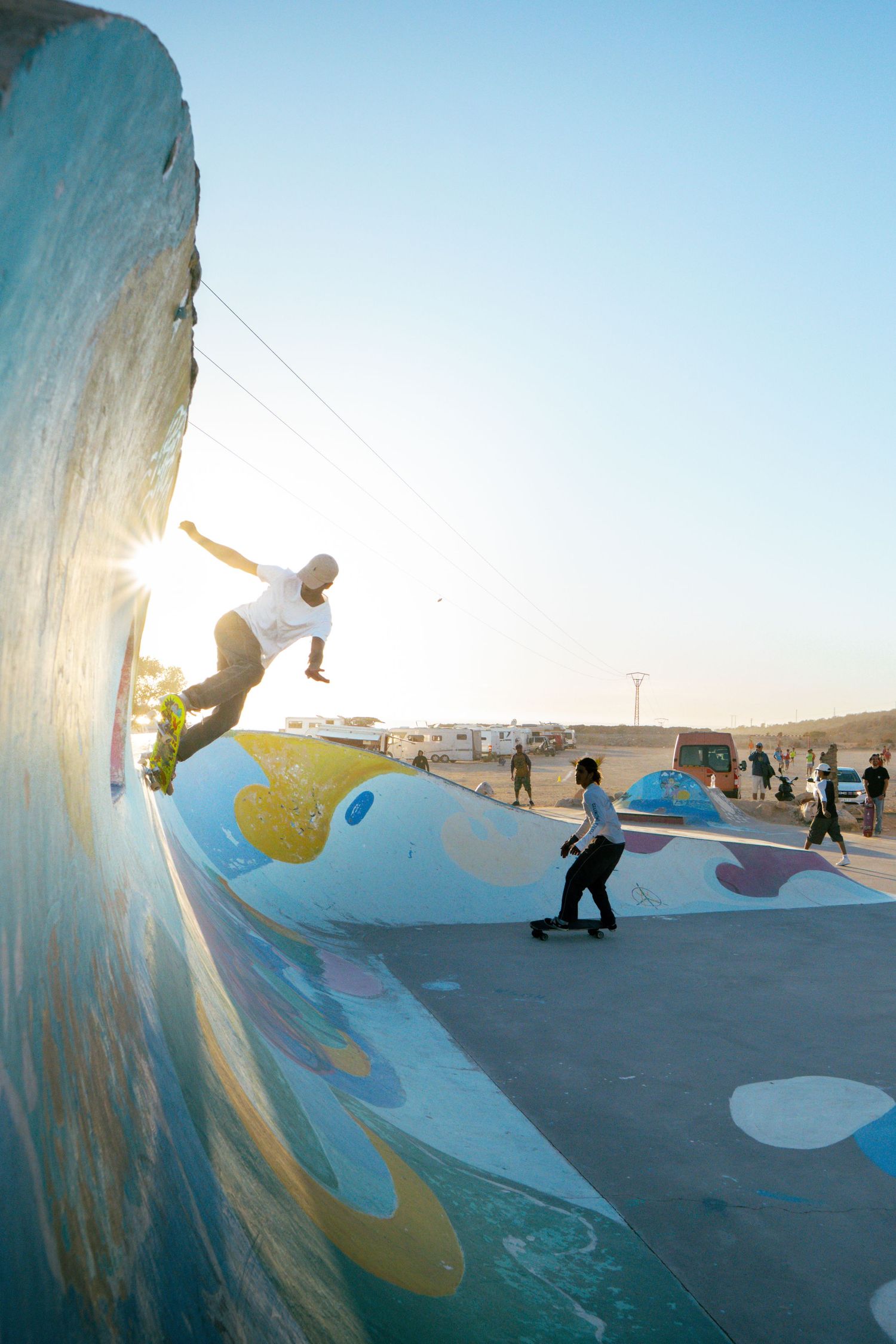 Skateboarder grinding wall of colorful painted skate ramp at sunset with lens flare at outdoor park.