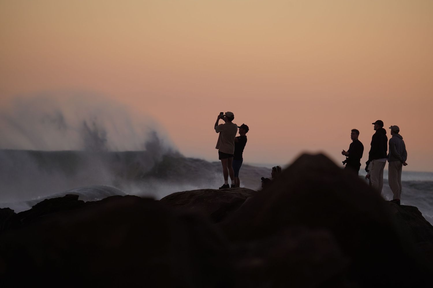 Silhouettes of people standing on rocky shore at sunset with dramatic ocean waves crashing in the background.