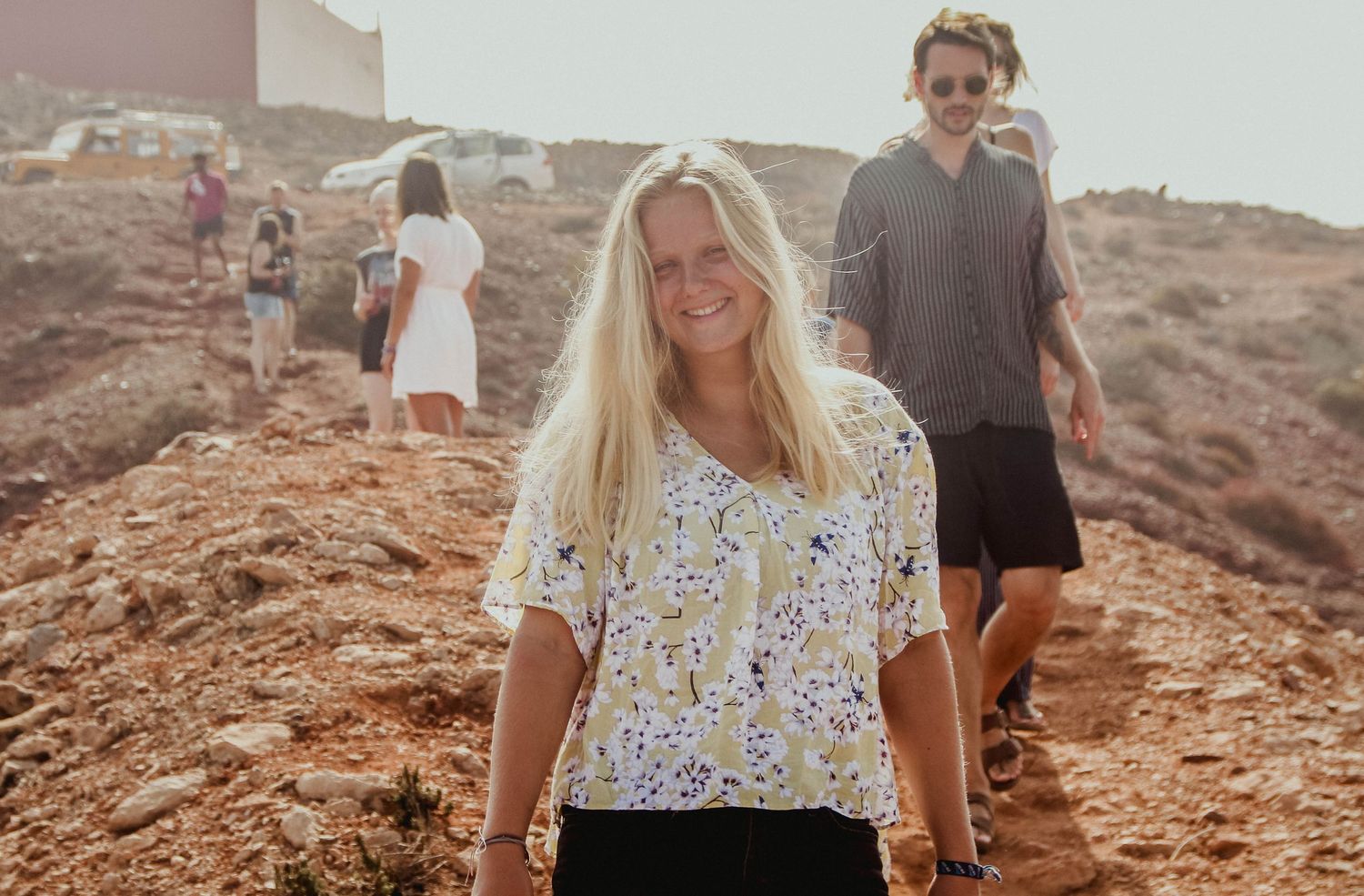 Smiling blonde woman in floral top on rocky terrain with people walking behind her in warm sunlight.