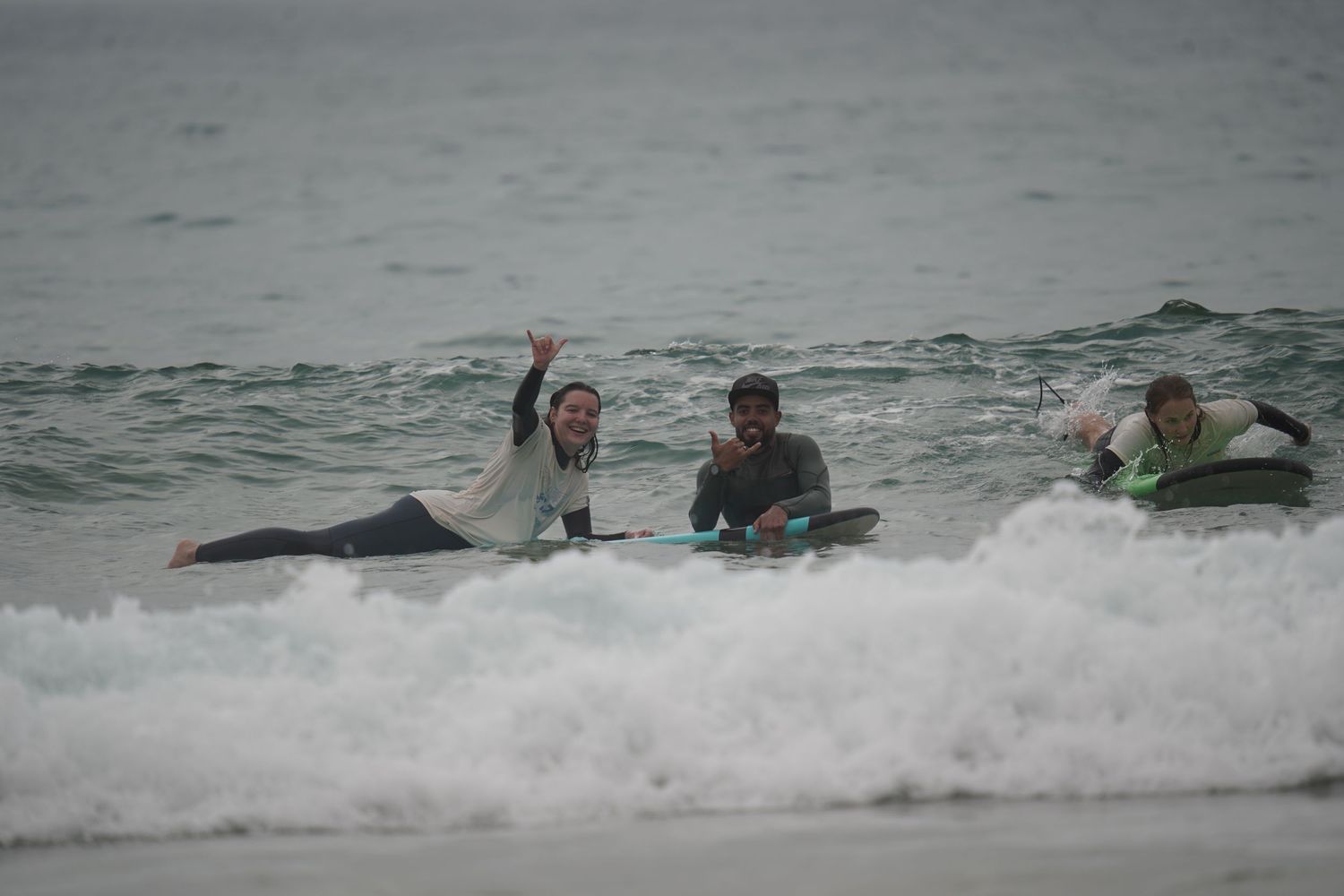 Three surfers riding waves together on a cloudy day, one raising their hand in celebration near the shoreline.
