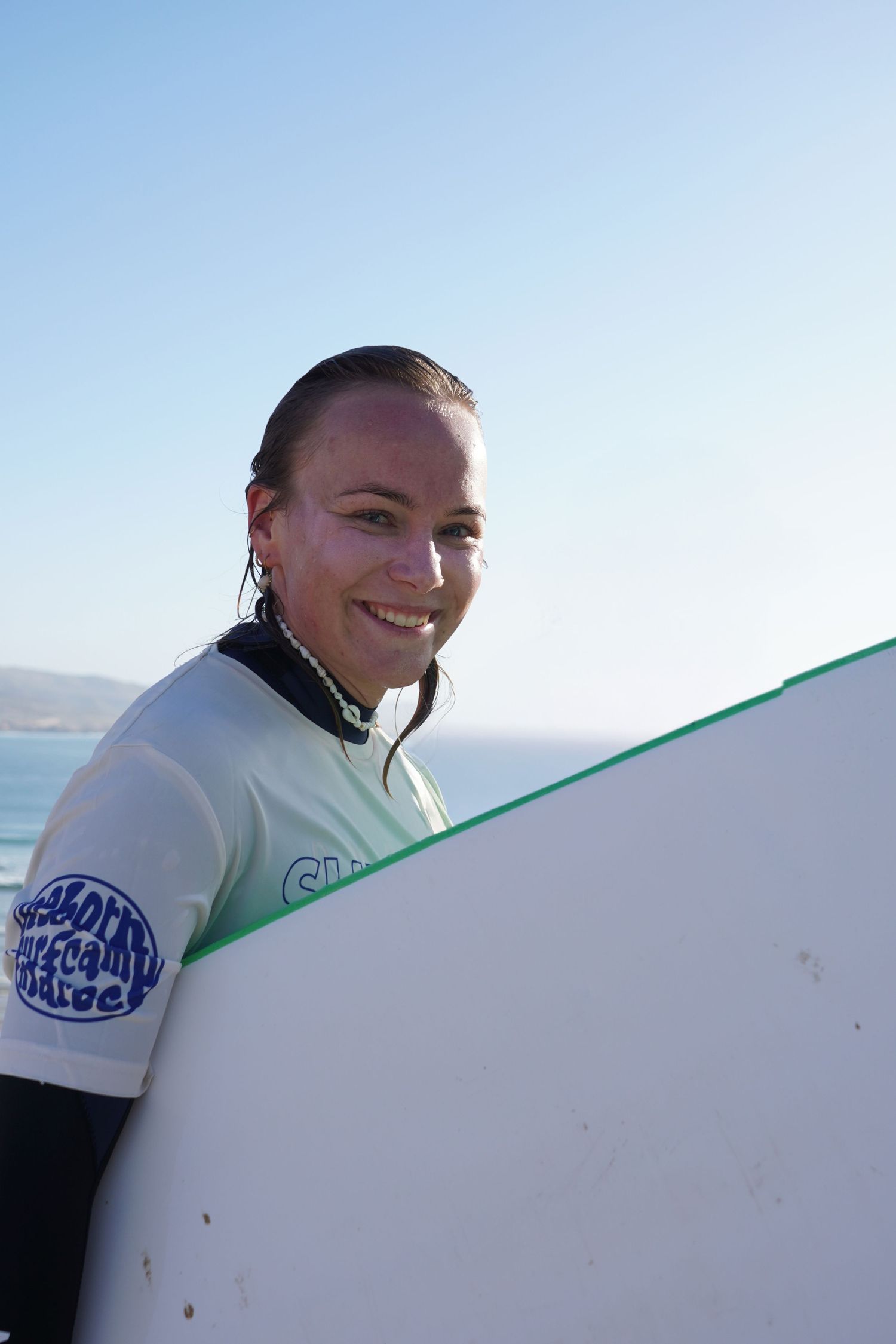 Smiling female surfer in wetsuit holding white surfboard on sunny beach with ocean in background.
