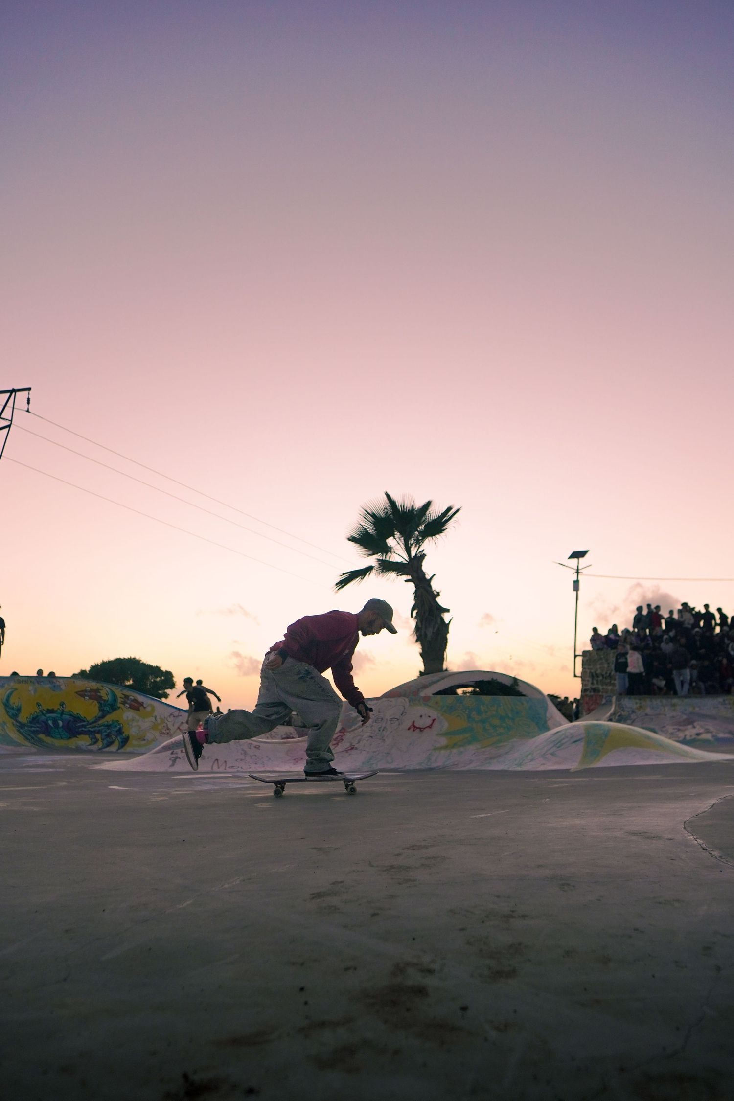 Silhouetted skateboarder performing trick at dusk with palm tree and purple sunset sky at skatepark.