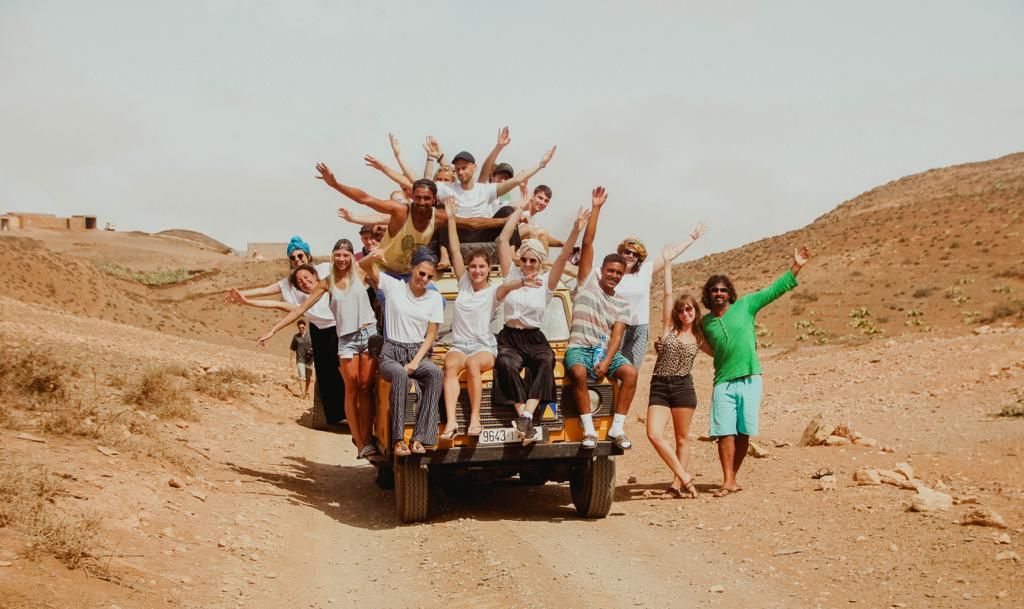 Group of excited young people riding on the back of a truck on a dusty desert road, waving and cheering.
