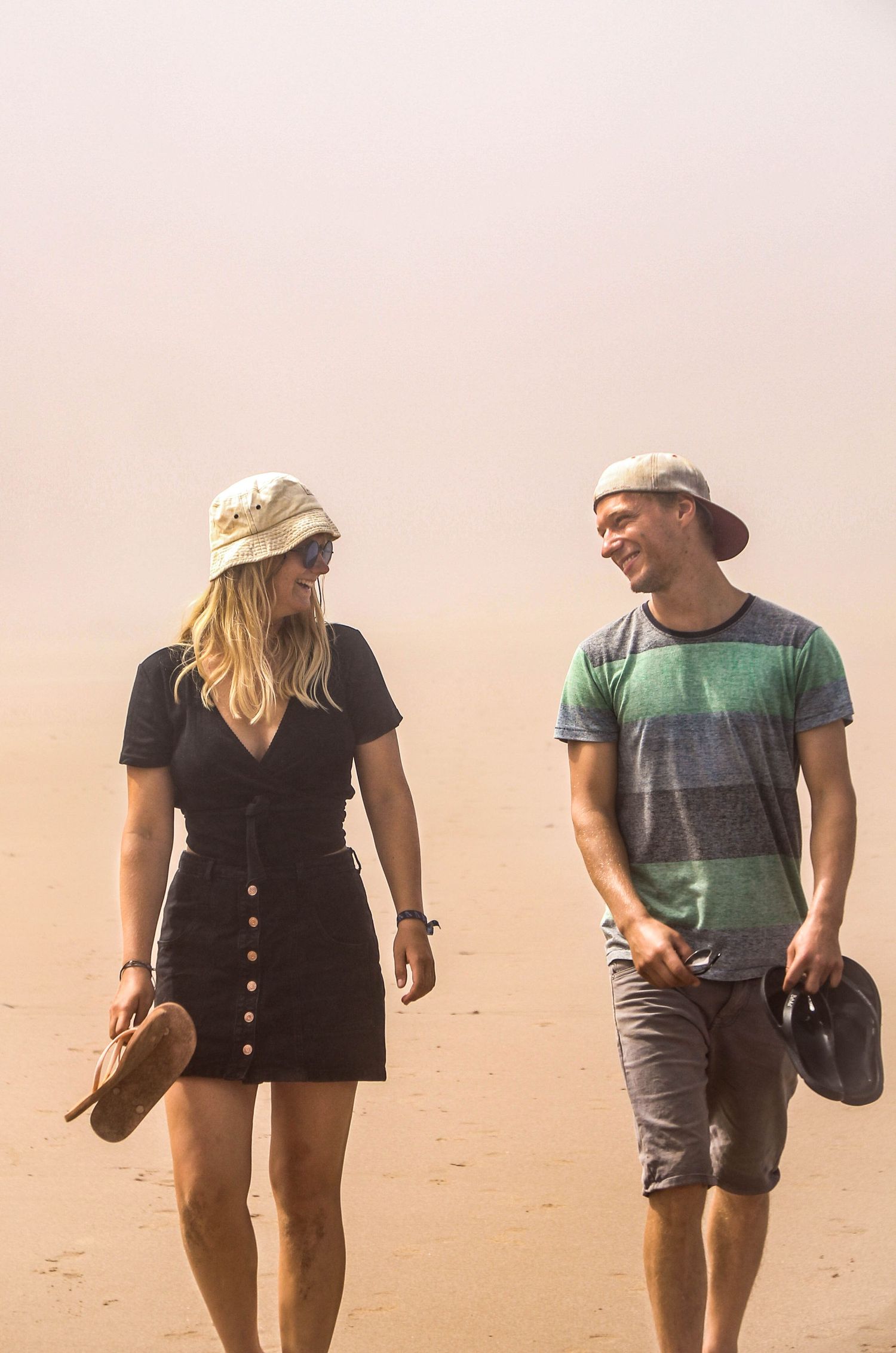A couple walks and smiles on a sandy beach, woman in black dress with hat, man in striped shirt holding a cap.
