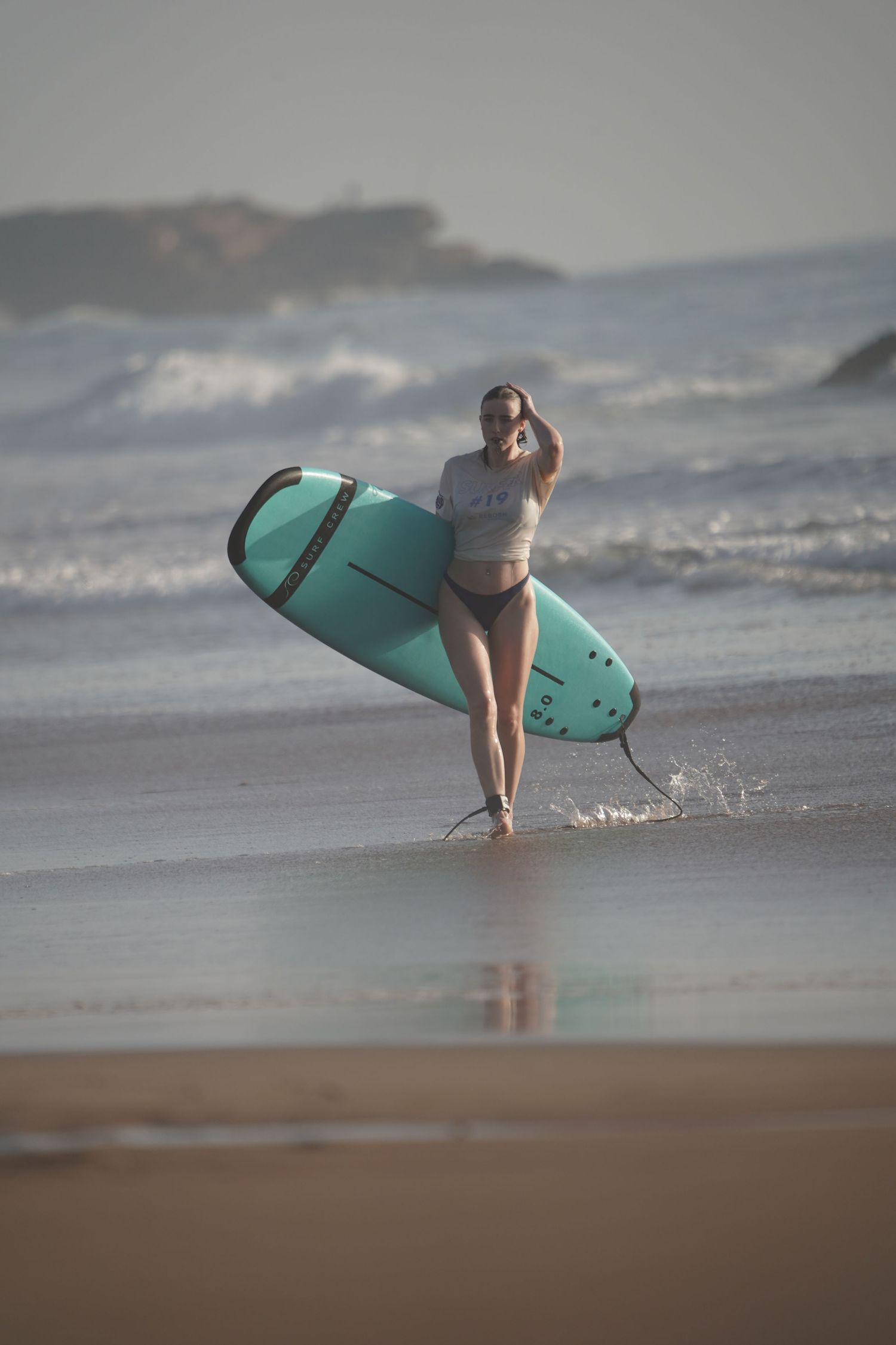 A woman in a bikini carries a turquoise surfboard while walking out of ocean waves on a sandy beach.