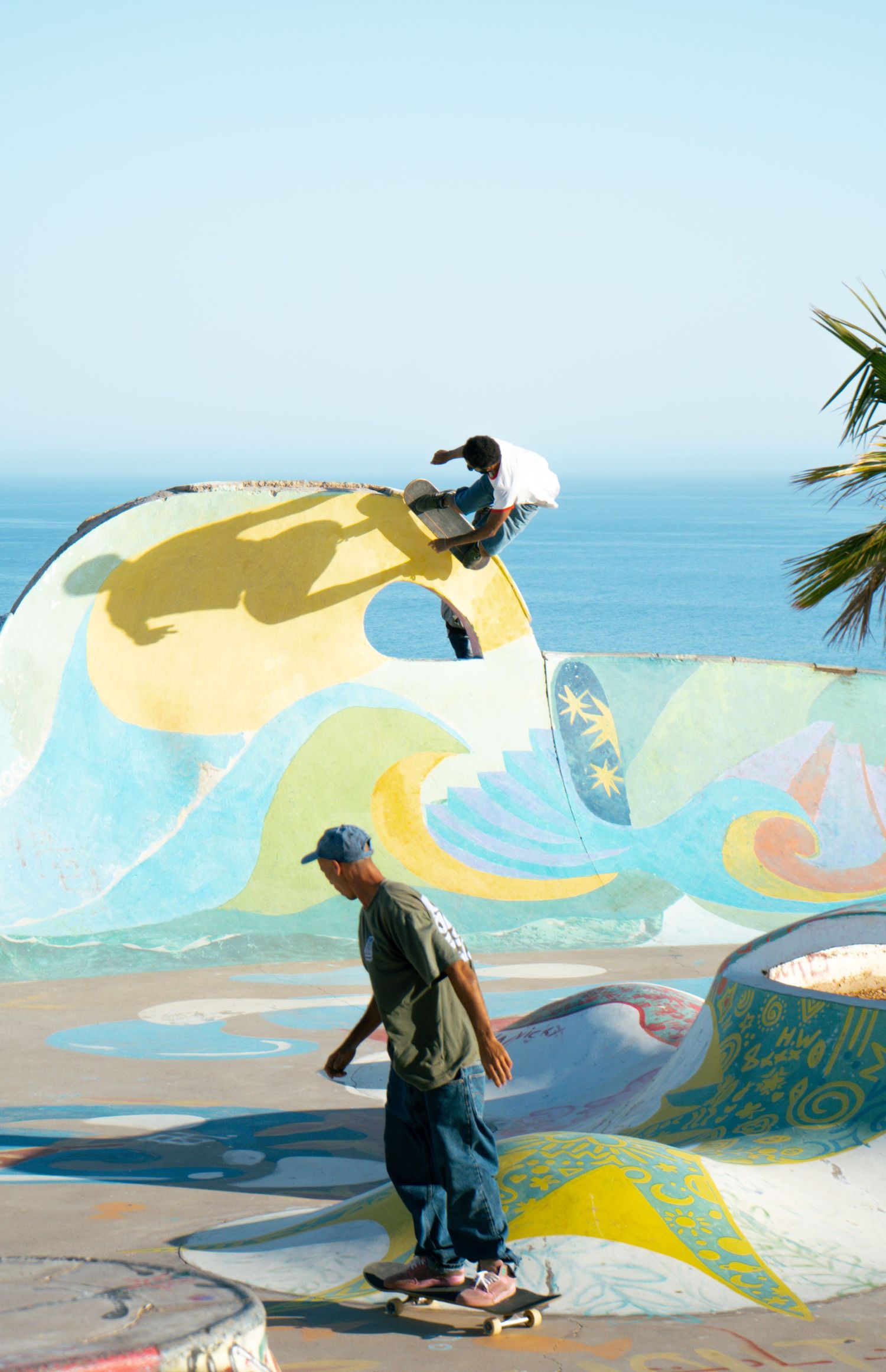 Two skateboarders ride colorful mural-painted ramps at oceanside skate park with palm tree and blue sea backdrop.
