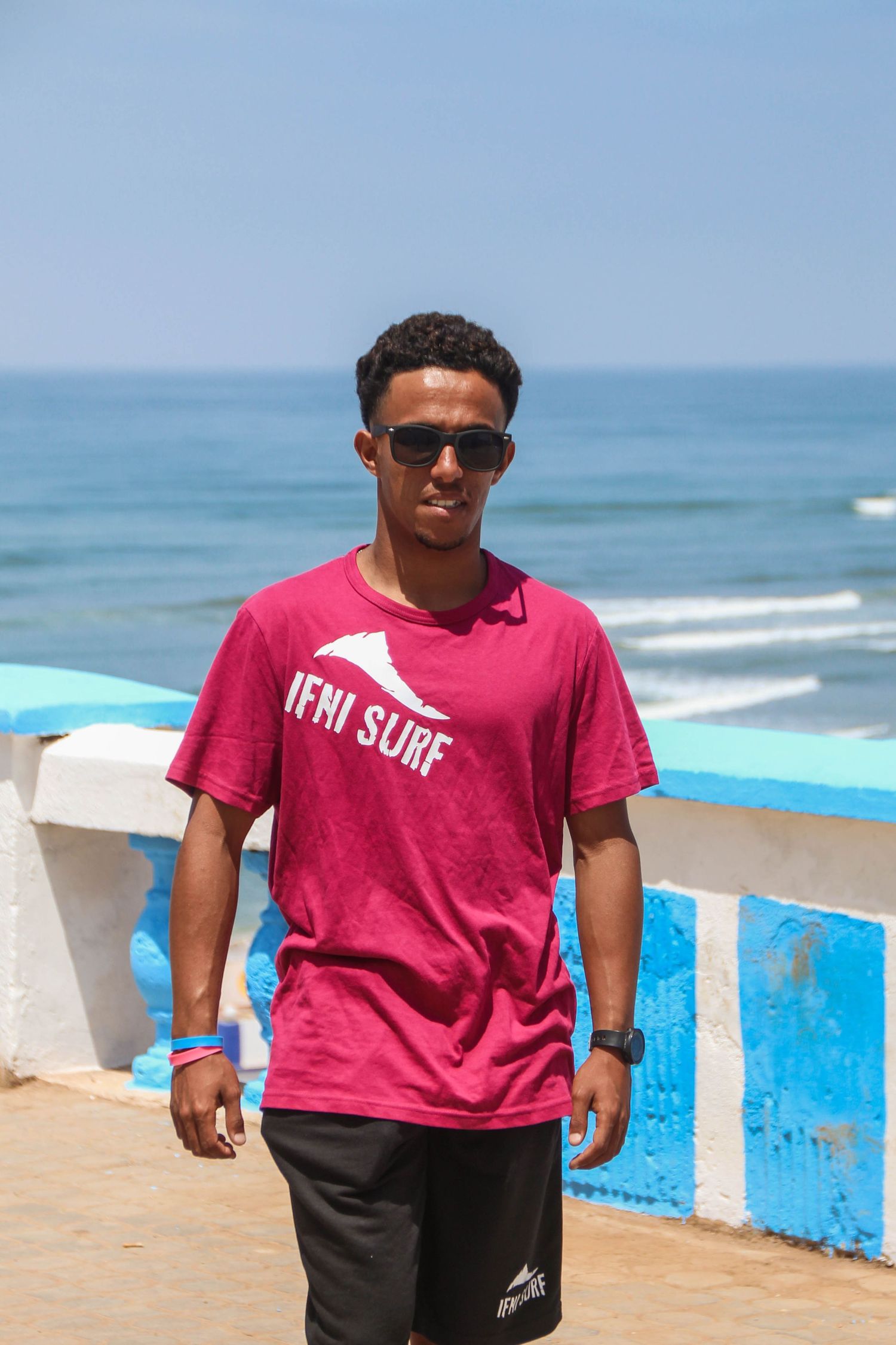 Young man in red Ifni Surf t-shirt and sunglasses standing on a seaside promenade with ocean waves behind him.
