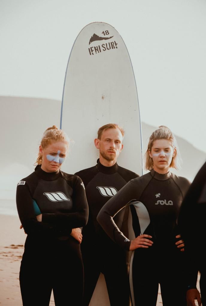 Three surfers in black wetsuits stand on a beach holding a white Ifni Surf board, looking serious.